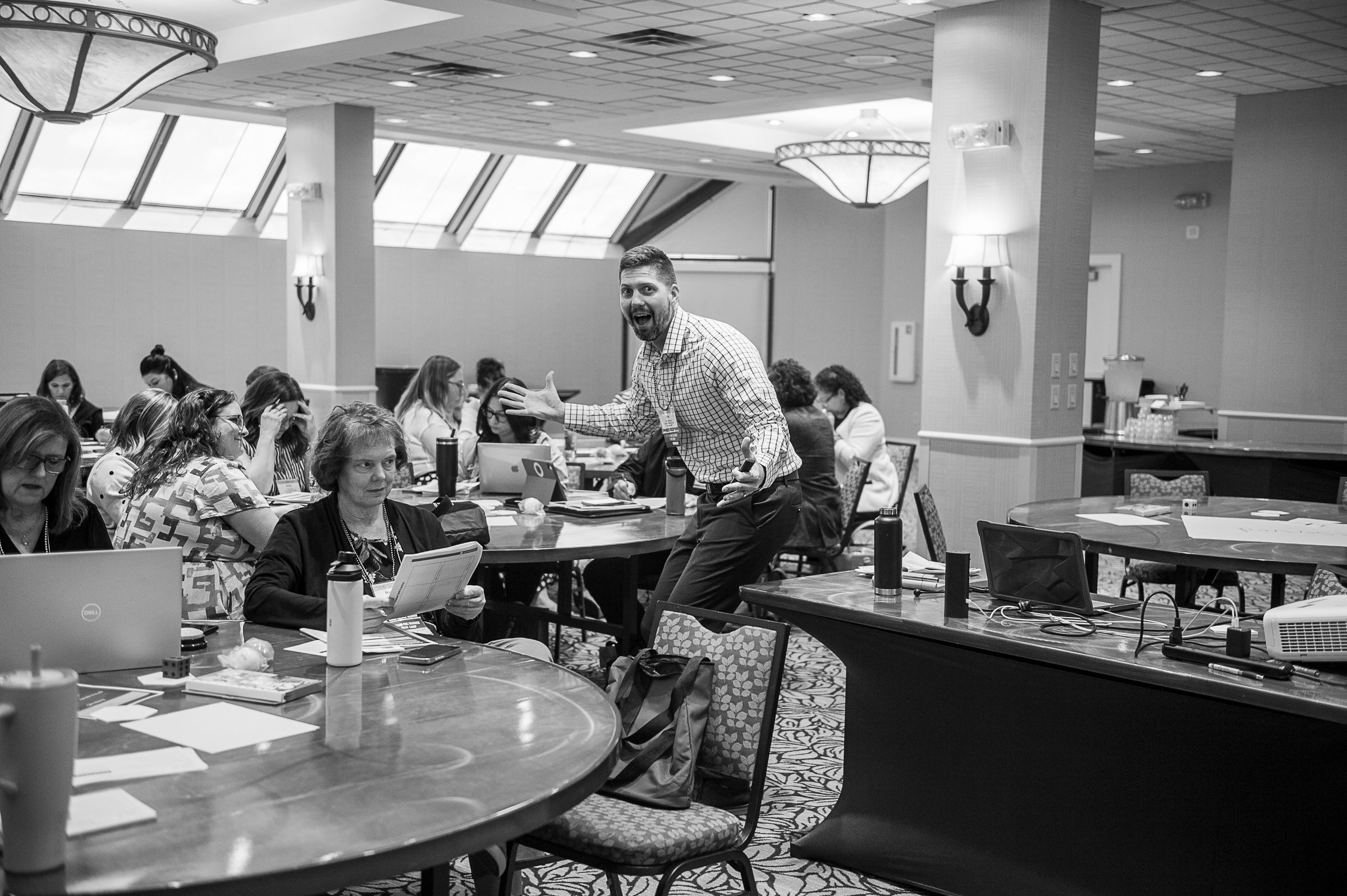 Tyler Moberg laughing and gesturing between round tables of participants during a training session. Black and white photo.