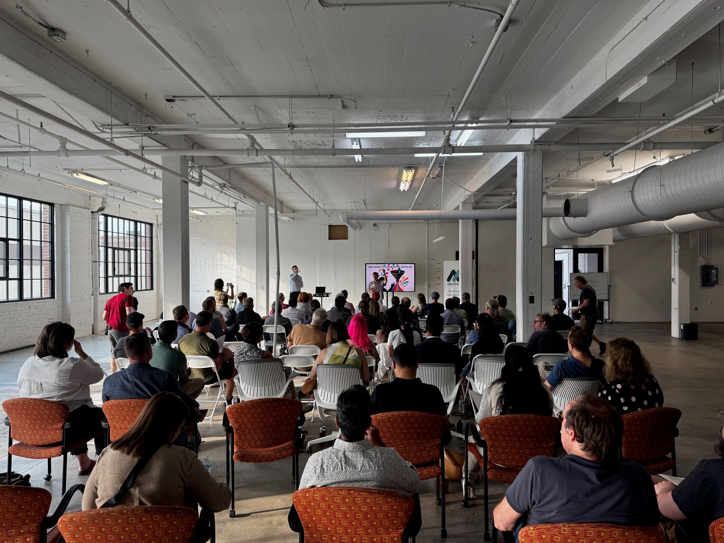 A full room seen from the back in a bright loft venue, participants facing the presenter and screen.