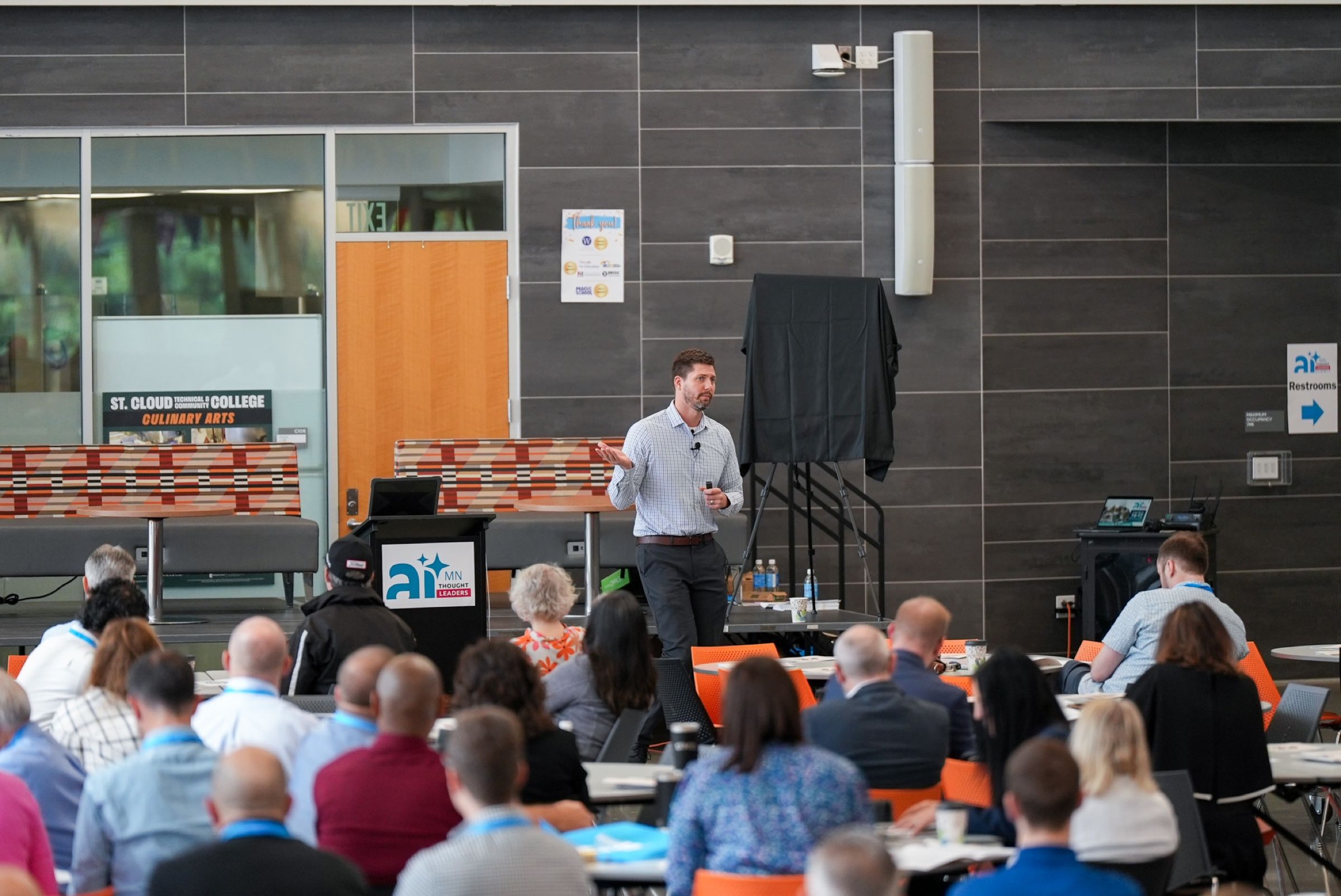 A summit room seen from the back, participants at round tables following along as Tyler teaches from the front.