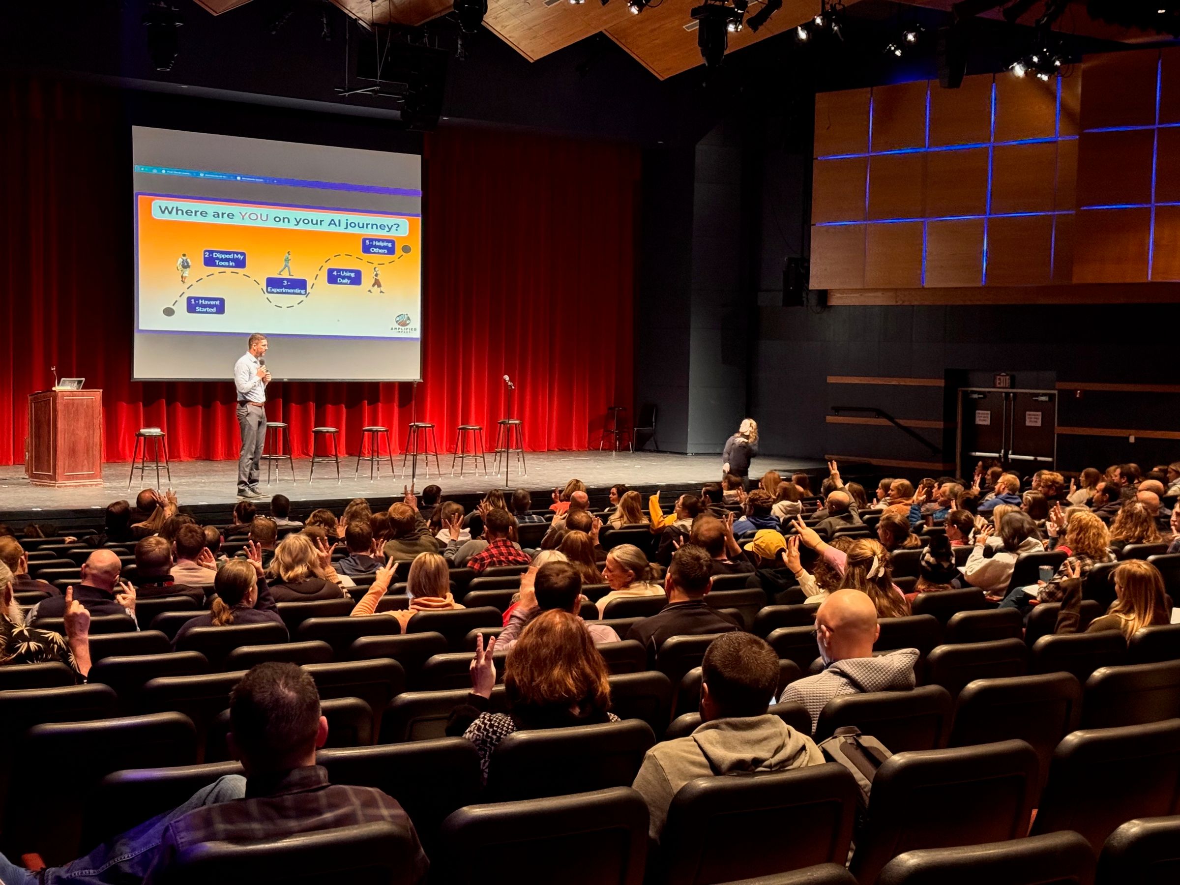 A full theater from the seats, hands raised across the room as Tyler asks a question from the stage.