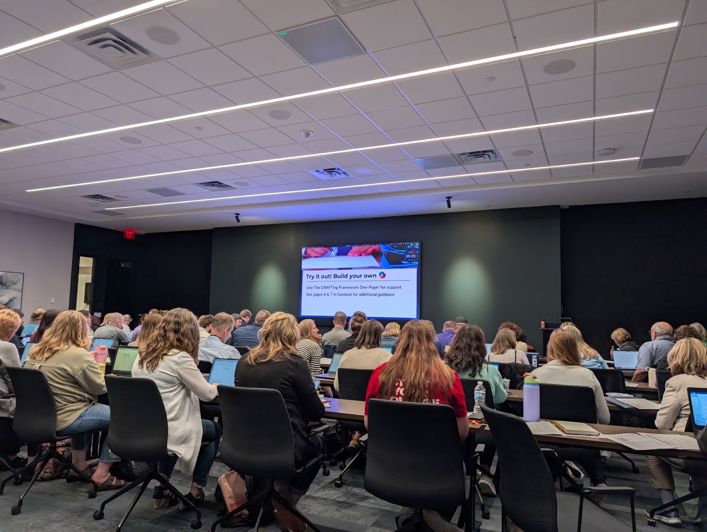 A training room from the back, participants on laptops at long tables facing a slide that reads Try it out! Build your own.
