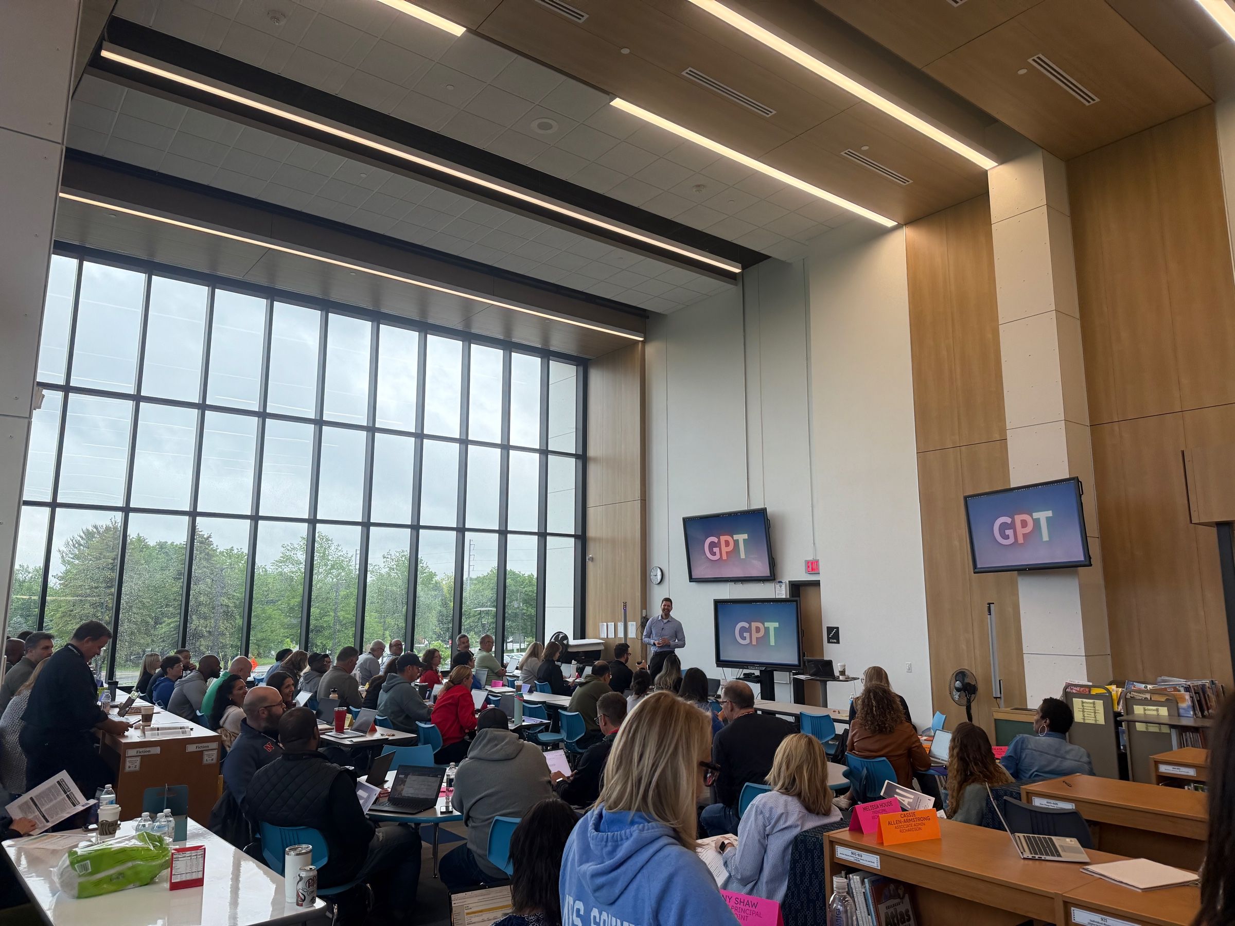 A large, sunlit hall full of participants at tables, GPT on the screens up front.