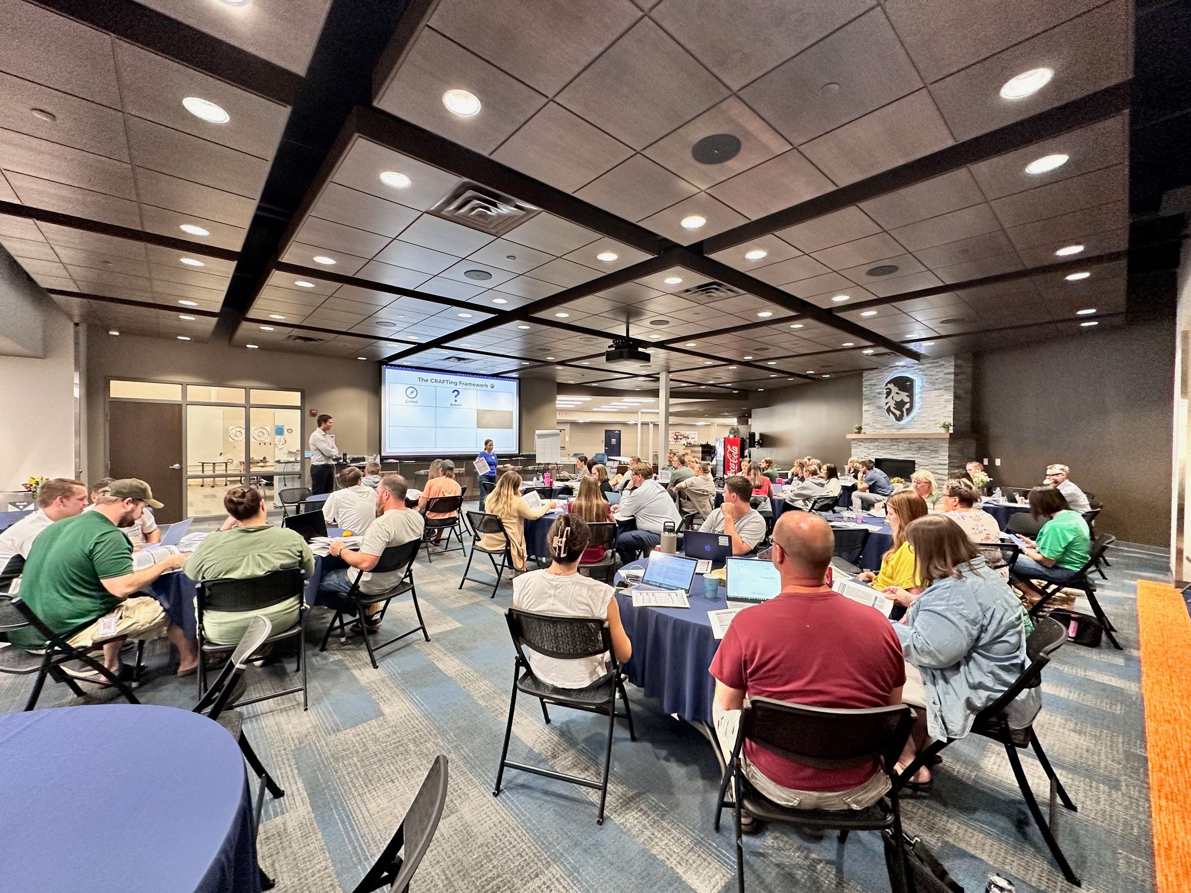 A full workshop room of round tables, participants working on laptops while Tyler teaches from the front.