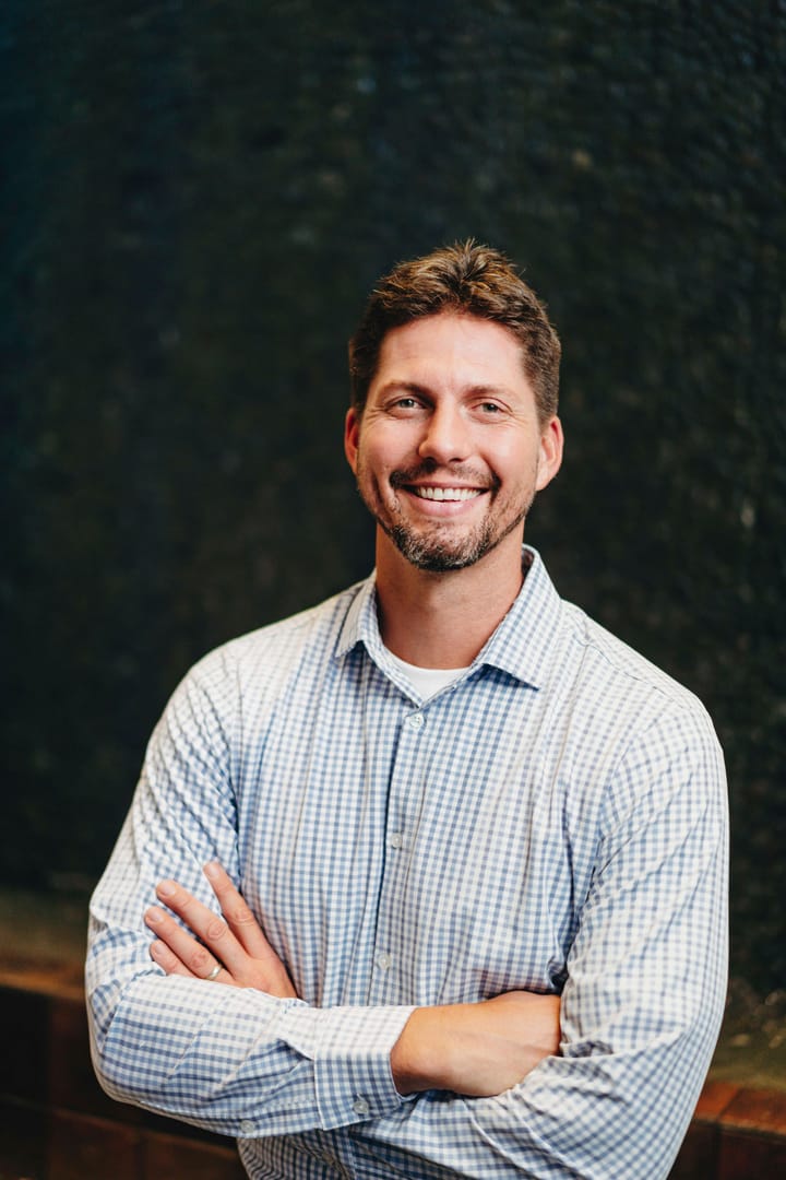 Tyler Moberg smiling with his arms crossed, in a checked shirt, against a dark textured wall.