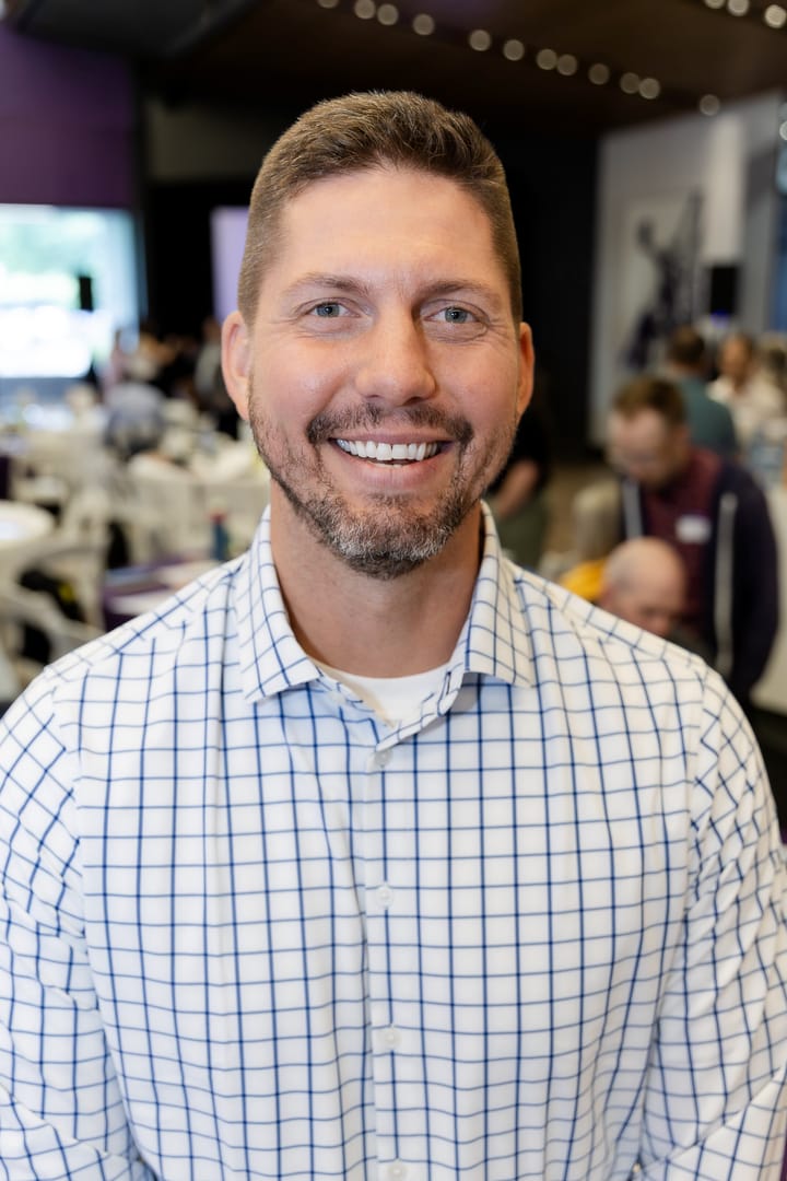 Headshot of Tyler Moberg smiling, in a blue checked shirt, with an event room behind him.