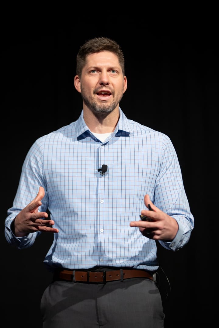 Tyler Moberg speaking on stage with a lapel mic, gesturing with both hands, against a black backdrop.